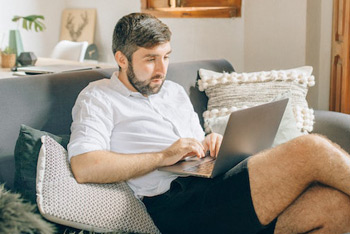 Mann im Homeoffice sitzt beim Arbeiten auf Sofa, Laptop auf den Knien.