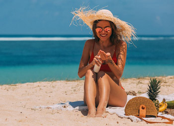 Junge Frau mit Sonnenhut cremt sich am Strand mit Sonnencreme ein