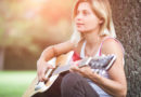 Junge Frau, die im Grünen an einen Baum gelehnt, Gitarre spielt.
