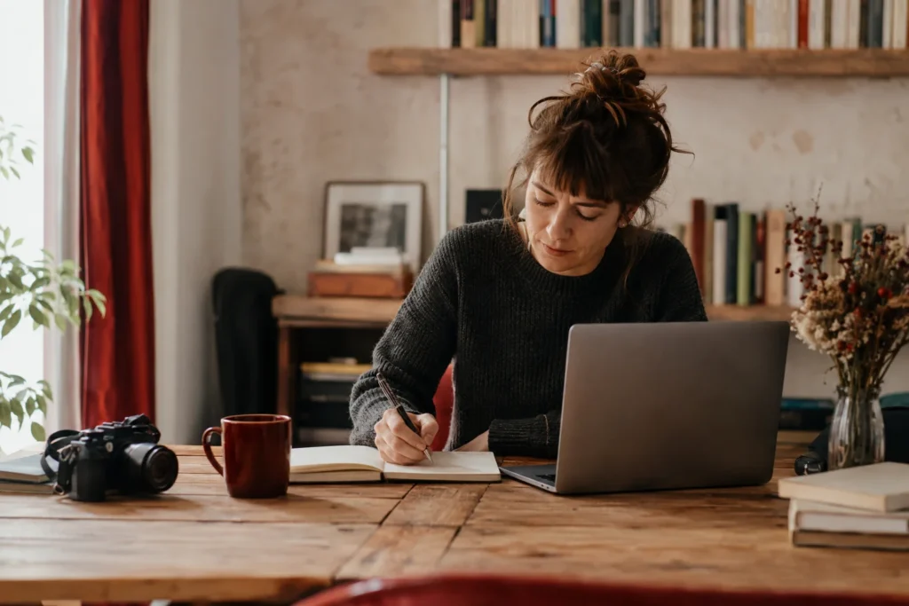 Bloggerin schreibt neben ihrem Laptop in ein Notizbuch; auf dem Tisch stehen eine Kamera und eine rote Tasse.